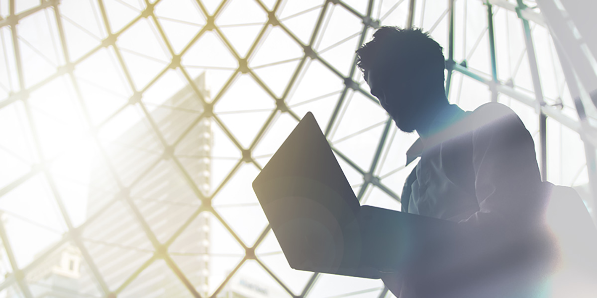 Business man walking with laptop and is silhouetted against a geometric glass window with a city skyline in the background